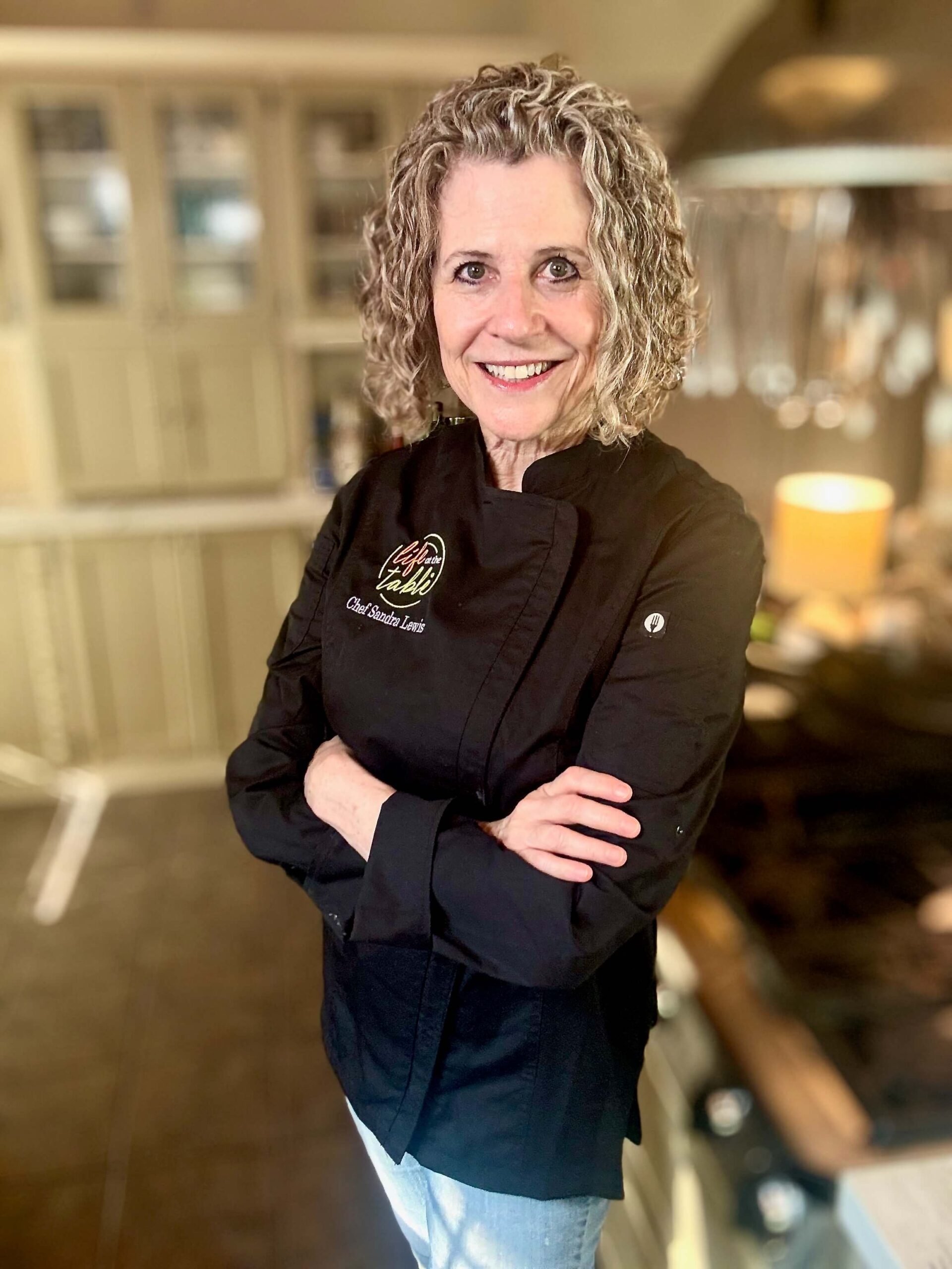 Chef Sandra Lewis Headshot 2 Chef Sandra Lewis Life At The Table in a black chef jacket standing in her kitchen with her arms crossed.